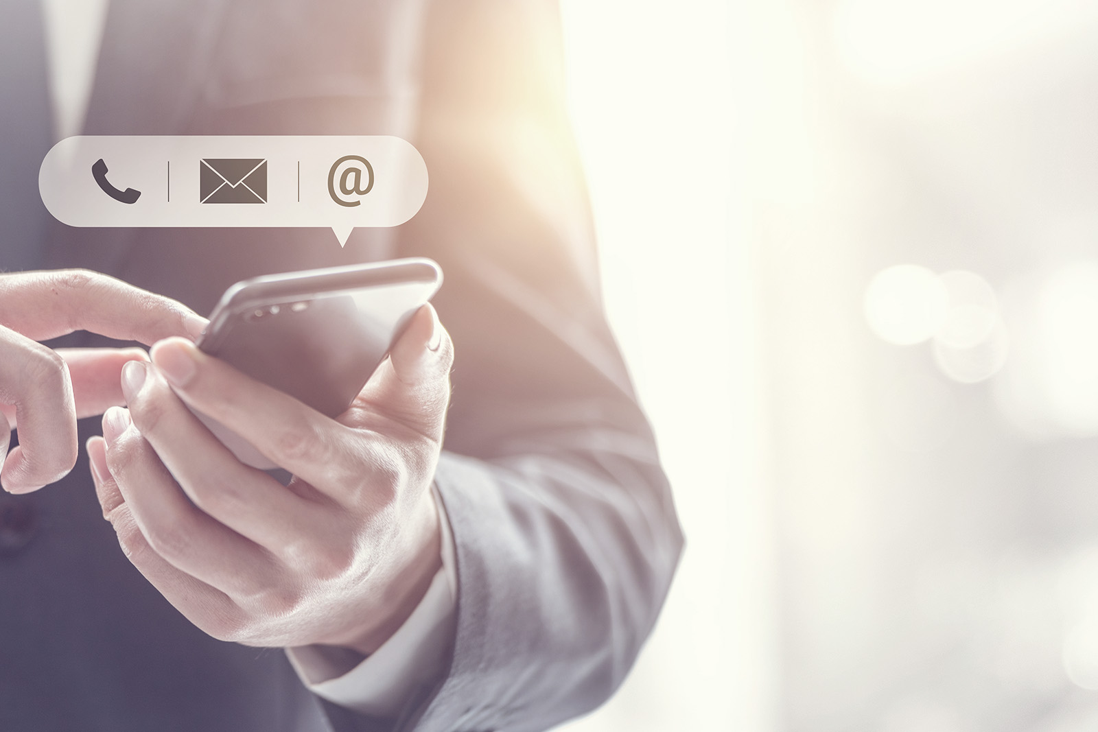 The image shows a close-up of a hand holding a smartphone, displaying an email icon on the screen. In the background, there is a blurred figure of a person in a suit, suggesting professional context, and the focus is on the phone s interface.